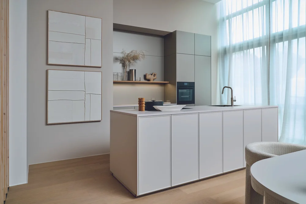 Strak keukeneiland in zachte greige tinten met een eiken vloer en lichtinval door een groot raam