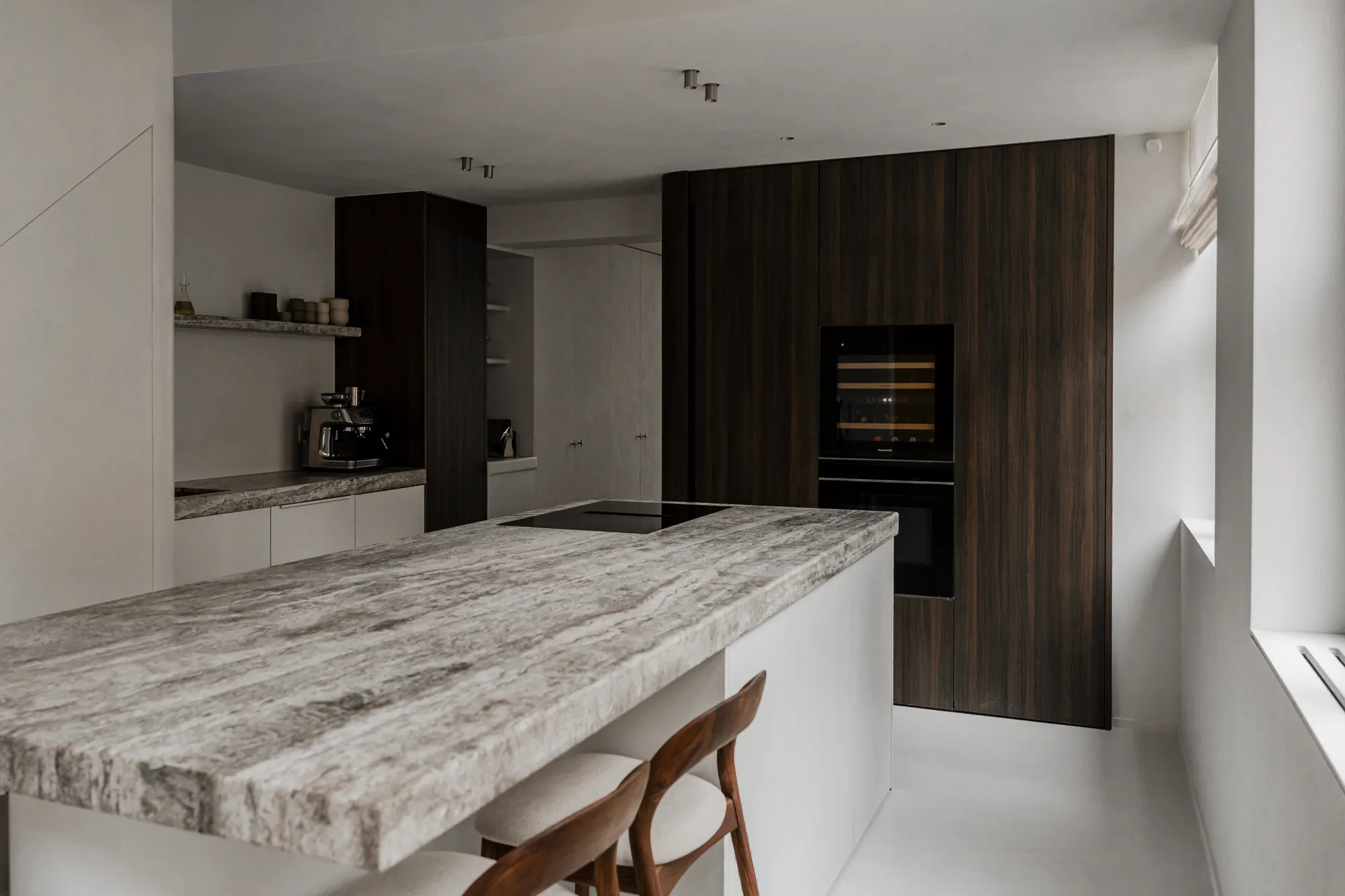 Keuken met natuurstenen eiland en kookplaat, een koffiehoek met zwevende plank links en een kastenwand in donker hout met wijnklimaatkast en oven